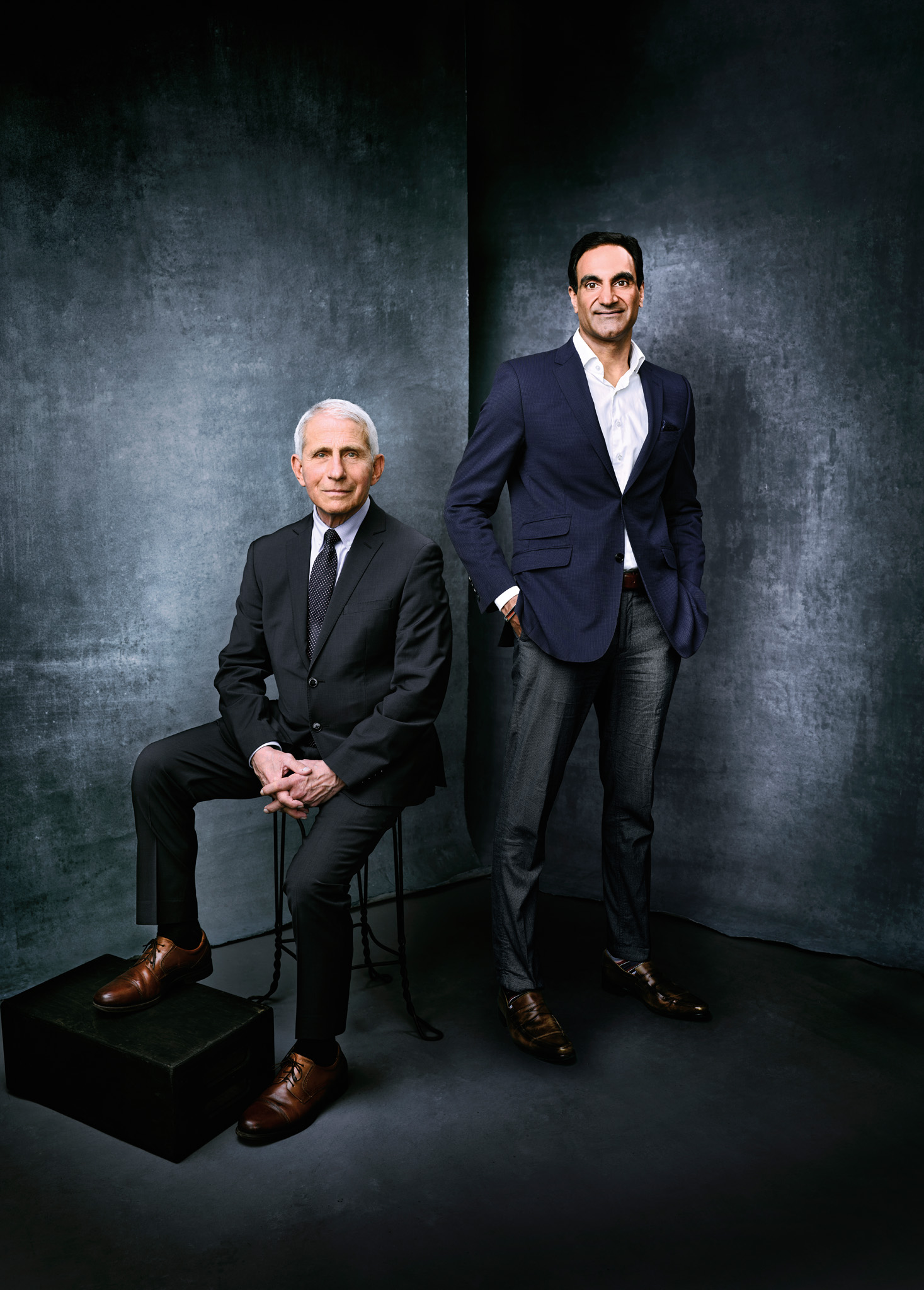 Older white man (left) wearing a powder blue collared shirt, navy blue suit and a navy tie with white dots, seated next to a tall South Asian man (right), in a white collared shirt, navy blue suit jacket and black pants.