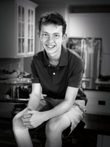 A 15-year-old white boy with short hair and freckles, wearing shorts and a dark colored, short-sleeved polo shirt, sitting on his kitchen counter. He is Chiari malformation patient Parker Riese.