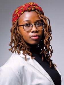 Portrait of a young Black woman wearing a white student doctor coat, circular glasses, and a red-patterned head scarf over her light orange-brown locs.