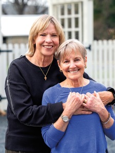From left: A blonde-haired middle aged white woman, wearing a black long sleeved shirt, hugs an older white woman, wearing a blue sweater, as they smile for a portrait