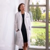 Vertical portrait of a middle aged Asian woman wearing a white coat over a black dress, with her hands in the pockets of her white coat