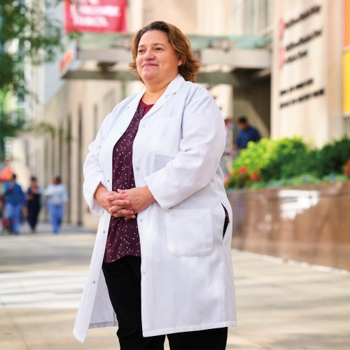 Latina woman in white coat with chin-length hair standing in front of limestone buildings with arched windows.