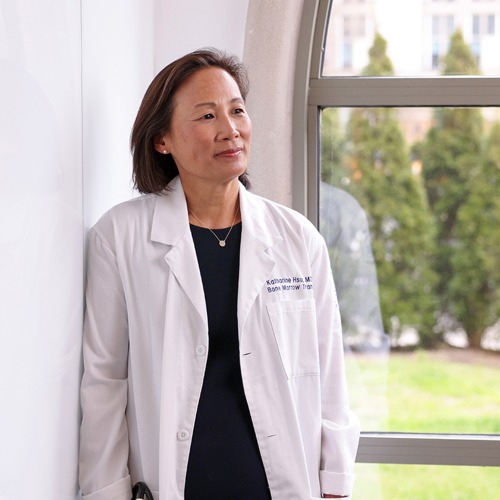 Vertical portrait of a middle-aged Asian woman wearing a white coat over a black dress, with her hands in the pockets of her white coat.