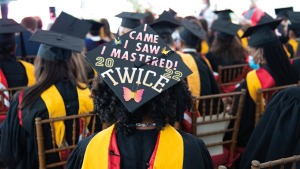 Graduates of Weill Cornell Graduate School of Medical Sciences are shown seated in black, yellow and red robes and black mortarboards from the back; mortarboard of person in foreground says, “I came, I saw, I mastered twice.”