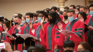 Diverse male and female graduates of Weill Cornell Medicine wearing red and green robes and face masks stand in rows with their right hands up at Commencement. 