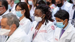 East Asian woman, Black woman in glasses and South Asian man, all in face masks and white coats, read materials during White Coat Ceremony for entering medical students.
