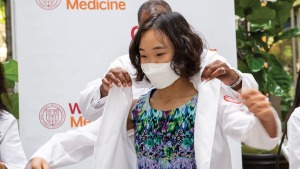 Dark skinned man puts short white coat on East Asian female medical student in white face mask.