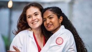 Two female medical students, one white with long brown hair, one South Asian with long, curly black hair, embrace and smile in their new white coats.