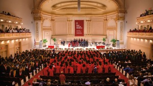 Medical college graduates seated in the audience facing a large stage, wearing red gowns and black caps during their commencement ceremony