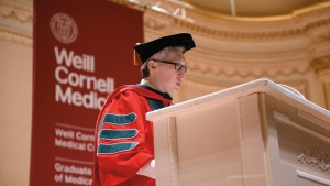 Middle aged Asian man in glasses, wearing a red gown and black cap, stands at the podium to address students