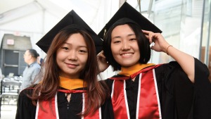 Two young Asian women, wearing red gowns and black caps, smile for a photo during a convocation ceremony