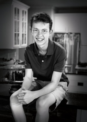 A 15-year-old white boy with short hair and freckles, wearing shorts and a dark colored, short-sleeved polo shirt, sitting on his kitchen counter. He is Chiari malformation patient Parker Riese.