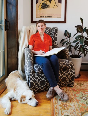 Tall, young white woman with short hair and slight smile, Dr. Laura Kolbe, assistant professor of medicine, sits wearing dark orange shirt, blue slacks and beige moccasins while holding large open book and pen in a home-like setting with Golden Retriever at her feet.