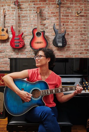 A Puerto Rican woman with curly hair, glasses and a pink blouse, sits in front of a piano as she looks to the side while she plays a blue guitar.
