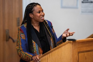 Candid photo of a Black woman speaking at a podium wearing a blue, yellow, red and black African printed blazer.